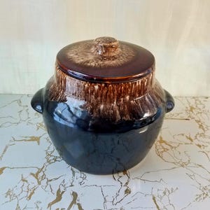 May include: A vintage ceramic bean pot with a lid. The pot is dark blue at the base, transitioning to a brown and tan glaze at the top. It has two small handles and a matching lid with a knob.