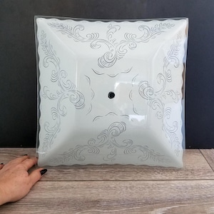 May include: Square, white ceiling light fixture with a decorative, etched scroll design. The fixture has a scalloped edge and a central black dot. The light is in a clear plastic wrap.