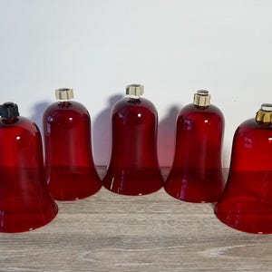 May include: Five red glass candle holders with silver and black accents. The bell-shaped holders are arranged on a wooden surface. The candle holders are a deep red color and have a glossy finish.