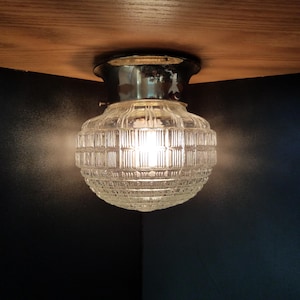 May include: A vintage-style ceiling light fixture with a clear, ribbed glass shade. The light is illuminated, casting a warm glow. The fixture has a chrome-colored base and is mounted on a wooden surface.