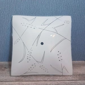 May include: Square, frosted glass light fixture with a wheat-like design in black. The fixture has a single, small, black circular hole in the center. The design is symmetrical and covers the entire surface of the glass.
