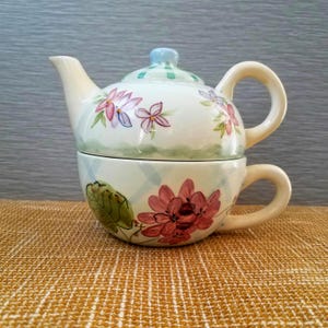 May include: A ceramic teapot and teacup set. The teapot is white with pink and purple floral designs and a light blue lid. The teacup is white with a pink flower design and a green leaf. The set is on a tan surface.