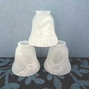 May include: A set of three bell-shaped glass lamp shades featuring a frosted, semi-translucent finish with a subtle, swirling white marble-like pattern throughout the surface. Each shade has a wide, flared opening at the bottom and a narrower neck.