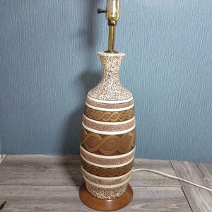 May include: A ceramic table lamp with a tall, bottle-shaped base featuring a speckled beige and brown finish. The body is decorated with horizontal bands of raised, interlocking brown rope patterns. It sits on a circular wooden base.