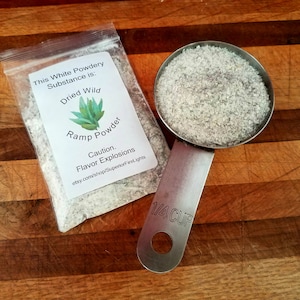May include: A clear plastic bag and a metal measuring cup filled with a white, powdery substance. The bag has a label that reads "Dried Wild Ramp Powder" with a green plant illustration and the text "Caution. Flavor Explosions."