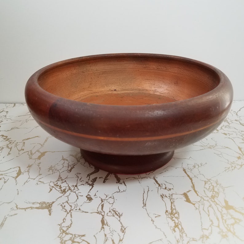 May include: A round, wooden bowl with a pedestal base. The bowl is a dark brown color with a lighter brown interior. A thin, lighter brown stripe encircles the bowl. The bowl is likely used for serving food or as a decorative item.