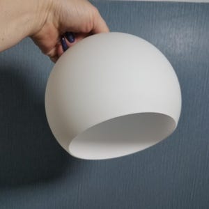 May include: A white, spherical lampshade with a smooth, matte finish. The lampshade has a curved opening at the bottom. The lampshade is being held by a hand against a textured blue background.
