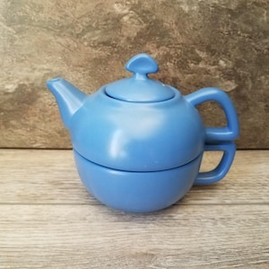 May include: A light blue ceramic teapot and teacup set. The teapot has a curved spout, a lid with a small handle, and a rounded body. The teacup is integrated into the base of the teapot, creating a compact design.