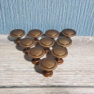 May include: A collection of ten bronze-colored cabinet knobs. Each knob has a round, flat top and a short, pedestal-style base. The knobs are arranged in a group, showcasing their design and finish. The hardware is suitable for furniture restoration or new builds.