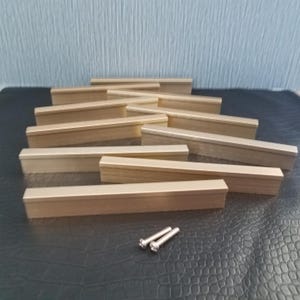 May include: A collection of rectangular, brass-colored cabinet pulls arranged on a dark surface. Two silver screws are visible near the bottom. The pulls have a simple, modern design, suitable for various furniture styles.