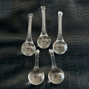 May include: Five clear glass teardrop-shaped ornaments. Each ornament has a round bulbous base and a long, slender stem that tapers to a small loop at the top. The ornaments are arranged on a textured black surface.