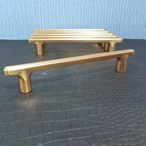 May include: Several gold-toned cabinet pulls with a T-shaped design are arranged on a dark surface. The pulls have a rectangular bar and rounded edges. The hardware is likely for kitchen or bathroom cabinets.