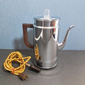 May include: A vintage chrome coffee percolator with a wooden handle and a clear plastic top. The coffee pot has a spout and is accompanied by a yellow and brown electrical cord with a plug. The percolator is a classic kitchen appliance.