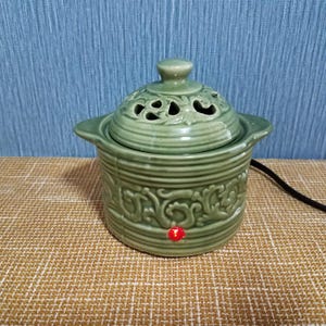 May include: A sage green ceramic electric wax warmer featuring a removable lid with decorative cutouts and a relief scroll pattern on the base. The cylindrical vessel includes two side handles and a small, circular red indicator light on the front.