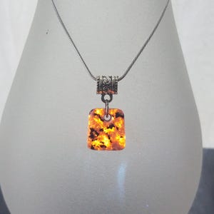 May include: A silver necklace with a rectangular pendant. The pendant is a mix of orange, yellow, and black, with a textured appearance. The necklace has a decorative silver clasp and a thin chain.