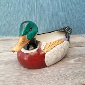 May include: A mallard duck-shaped ceramic bowl featuring a glossy green head, a yellow-brown beak, a dark red chest, and white and tan textured feather details on the body. The interior of the bowl is white, and the tail feathers are black.