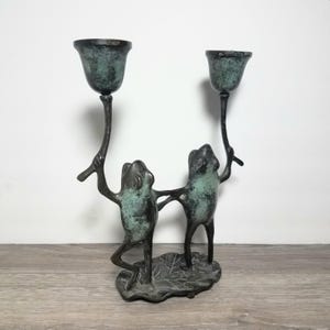 May include: A dual candle holder crafted from dark metal with a weathered green patina finish. The base is shaped like a lily pad, supporting two upright frog figures that each hold a tall, slender stem topped with a bell-shaped candle cup.