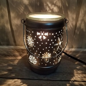 Puede incluir: Una linterna de metal marrón oscuro con recortes de copos de nieve. La linterna tiene una parte superior de vidrio y un asa de metal. La luz interior brilla a través de los diseños de copos de nieve, creando un brillo cálido. La linterna está sobre una superficie de madera.
