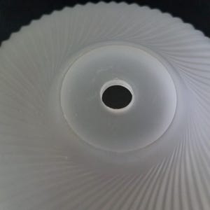 Może przedstawiać: Klosz lampy ze szkła matowego z spiralnym wzorem. Środek ma okrągły otw&oacute;r. Powierzchnia klosza ma teksturowany, żebrowany wz&oacute;r, kt&oacute;ry promieniuje na zewnątrz od środka. Og&oacute;lny kształt jest okrągły.