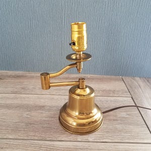 May include: A brass-toned, adjustable desk lamp with a bell-shaped base and a pivoting arm. The lamp features a standard light socket and a small on/off switch. A brown power cord extends from the base.