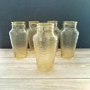 May include: Six amber-colored glass vases with a textured, ribbed design. The vases are cylindrical with a slightly flared top. They are arranged on a wooden surface against a dark blue background.