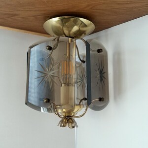 May include: A vintage ceiling light fixture with a brass finish and a clear glass shade. The shade is etched with starburst patterns. The light bulb is visible inside the fixture. The fixture is mounted on a wooden surface.