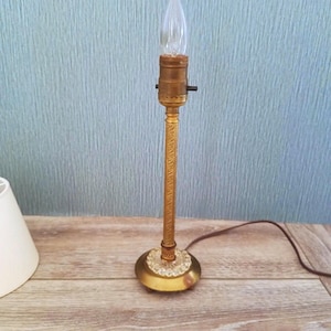 May include: A vintage brass table lamp with a decorative etched stem and a clear, pointed light bulb. The lamp has a round base with a crystal-like trim. A white lampshade is partially visible on the left.