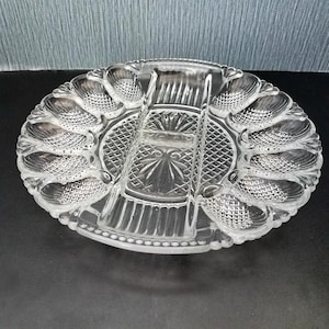 May include: Clear glass serving platter with multiple compartments. The oval-shaped dish features a central section and surrounding compartments with a textured, shell-like design. The platter is designed for serving appetizers or snacks.
