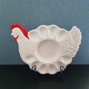 May include: White ceramic egg holder in the shape of a hen. The hen has a red comb and wattle, with a circular center and egg-shaped indentations. The holder is on a black metal stand.