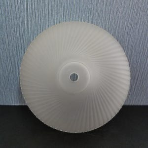 Może przedstawiać: Klosz lampy ze szkła matowego ze spiralnym wzorem. Klosz jest okrągły z centralnym otworem i teksturowaną powierzchnią. Kolor jest miękki biały, a wz&oacute;r promieniuje ze środka.