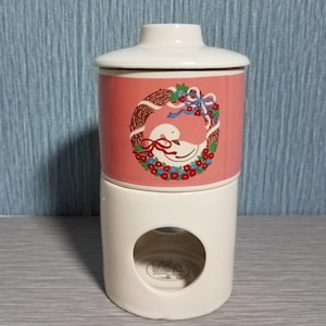 May include: A two-piece ceramic candle warmer with a white base and a pink upper section. The pink section features a wreath design with a white swan. The warmer has a lid and a circular opening on the base.