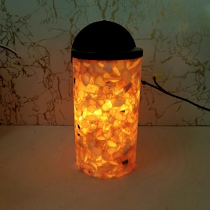 May include: A cylindrical lamp with a black top and base, filled with illuminated, irregularly shaped amber-colored stones. The lamp emits a warm, inviting glow, creating a cozy ambiance. The lamp is a decorative lighting fixture.
