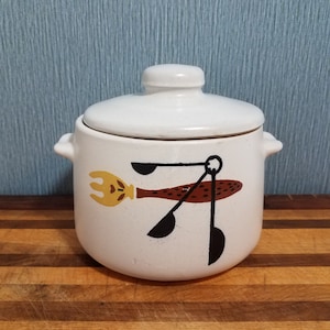 May include: A white ceramic casserole dish with a lid and two small handles. The dish is decorated with a brown and yellow fork and spoon design. The dish is sitting on a wooden cutting board.