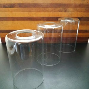 May include: Three clear glass lamp shades with a cylindrical shape and open tops. The shades are arranged in a row, with the closest one in focus. The background features a wooden cutting board.