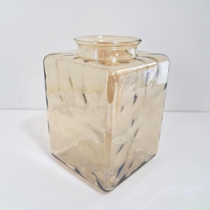 May include: A square, iridescent glass vase with a textured surface. The vase has a wide, open top and a warm, golden-brown color. The glass has a rippled effect, adding visual interest to the design.
