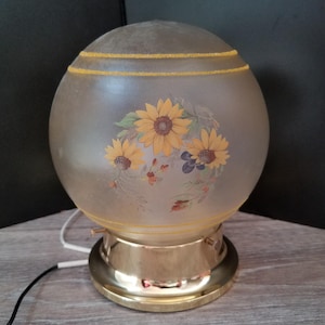 May include: A vintage globe lamp with a frosted glass shade. The shade features a floral design of yellow sunflowers and green leaves. The base is made of gold-colored metal. The lamp has a power cord.