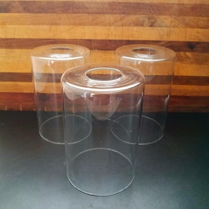 May include: Three clear glass cylindrical shades with a circular opening at the top. The shades are transparent, allowing light to pass through. They are likely for a lamp or light fixture.