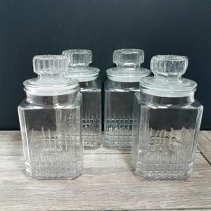 May include: A set of four clear glass canisters with square bases and matching glass lids. Each canister features a textured grid pattern on the lower half of the body and a fluted, knob-style handle on the lid. The containers are transparent.
