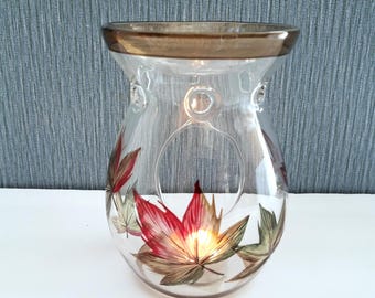 May include: A clear glass candle holder with a brown rim and a decorative leaf design. The leaves are painted in shades of red, green, and brown. The candle holder has a circular opening for a tea light.