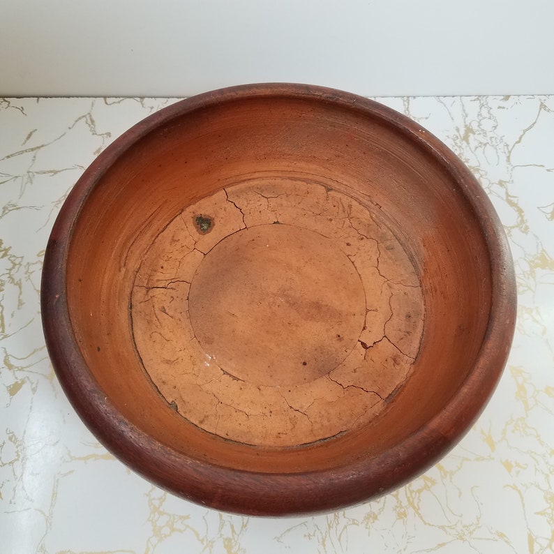 May include: A round, unglazed terracotta bowl with a dark brown rim. The interior of the bowl has a cracked, aged appearance. The bowl is empty and sits on a white and gold patterned surface. The bowl is likely for decorative or functional use.