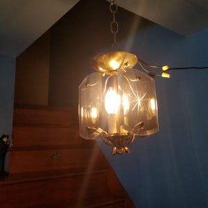 May include: Vintage brass and glass chandelier with starburst etched glass panels. The light fixture hangs from a chain and features three candelabra-style bulbs. The chandelier is suspended in front of a wooden staircase.
