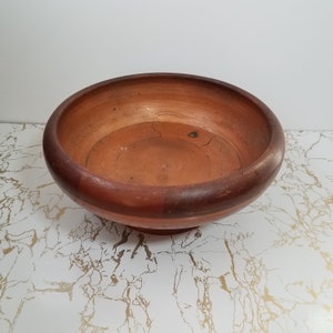 May include: A round, terracotta-colored bowl with a wide rim and a slightly raised base. The bowl has a rustic appearance with visible cracks and variations in color, suggesting a handmade or aged quality. The interior is a lighter shade of terracotta.