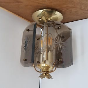 May include: A vintage ceiling light fixture with a brass finish and a smoky gray glass shade. The shade features etched starburst designs. The fixture has three light bulbs and is mounted on a wooden surface.