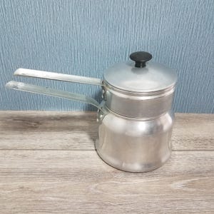May include: A silver-colored aluminum double boiler pot with a rounded base and a matching lid. The pot features two long, flat metal handles extending from the sides, and the lid is topped with a small, round, black plastic knob for handling.