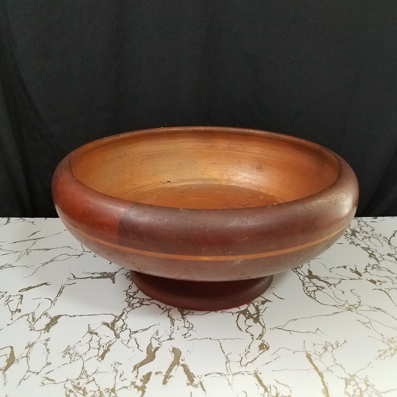 May include: A round, wooden bowl with a pedestal base. The bowl is a deep reddish-brown color with a lighter brown interior. A thin, gold-colored band encircles the bowl. The bowl is sitting on a white and gray marble-patterned surface.