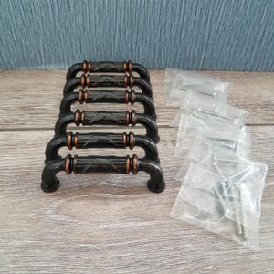 May include: A set of six black cabinet handles with copper-colored accents. Each handle has a decorative diamond pattern and comes with screws in individual plastic bags. The handles are arranged on a light wood-grain surface.