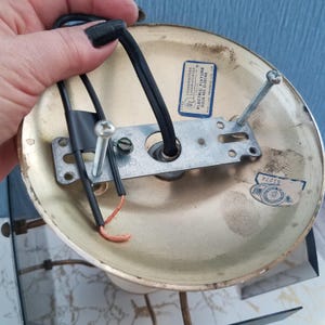 May include: A vintage brass-colored electric fixture base with a black wire and exposed copper wiring. The base has a metal bracket with screw holes and a UL label. The fixture is designed for electrical wiring and mounting.