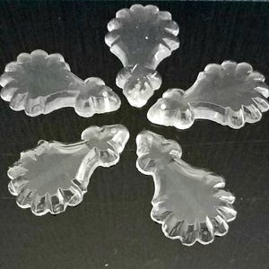 May include: Five clear, decorative crystal chandelier pieces arranged on a dark surface. Each piece has a scalloped edge and a stylized, curved shape, reflecting light. The crystals are likely for home decor.