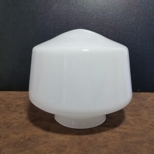 May include: A white, opaque glass lampshade with a rounded top and a flared, conical body. The lampshade has a smooth, glossy finish and sits on a small, circular base. The background is a dark brown surface and a black wall.