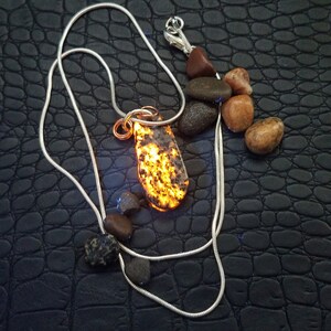 May include: A silver chain necklace with a pendant made of a glowing, speckled stone. The pendant is teardrop-shaped and has a copper wire bail. Several small, smooth stones of various colors are scattered around the necklace.